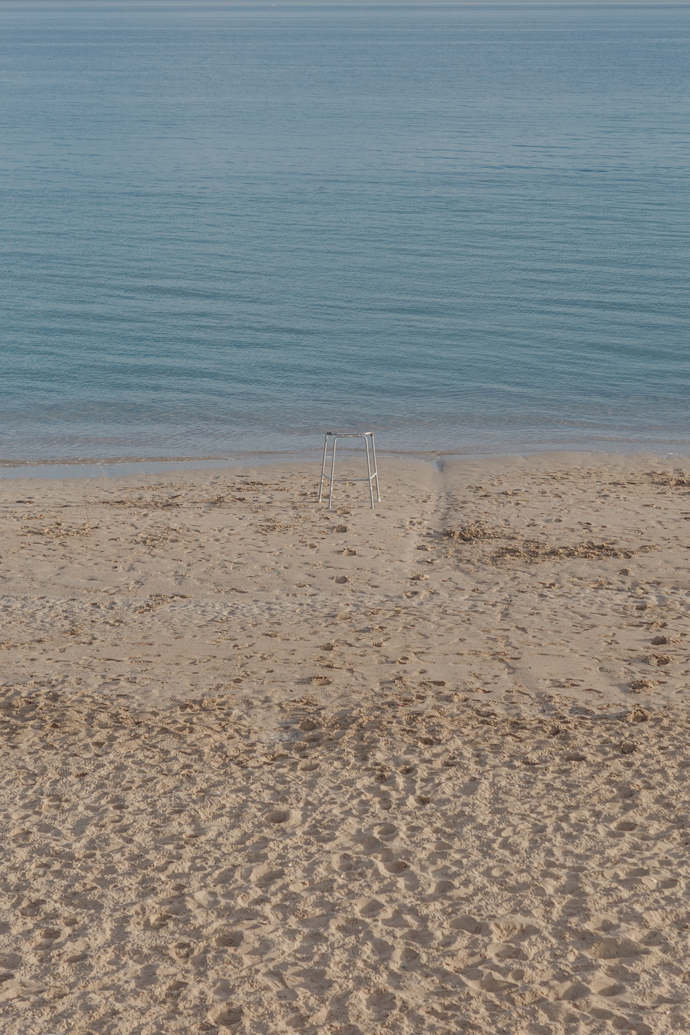 A sandy beach leading into calm blue waters with a lone metal walking frame partially submerged at the water's edge. Footprints are visible in the sand.