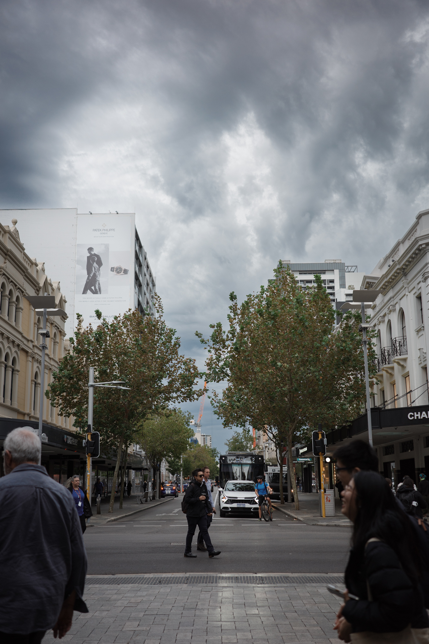Auto-generated description: A cloudy sky looms over an urban street scene with pedestrians, trees, and classic architecture intermingling with modern signage.