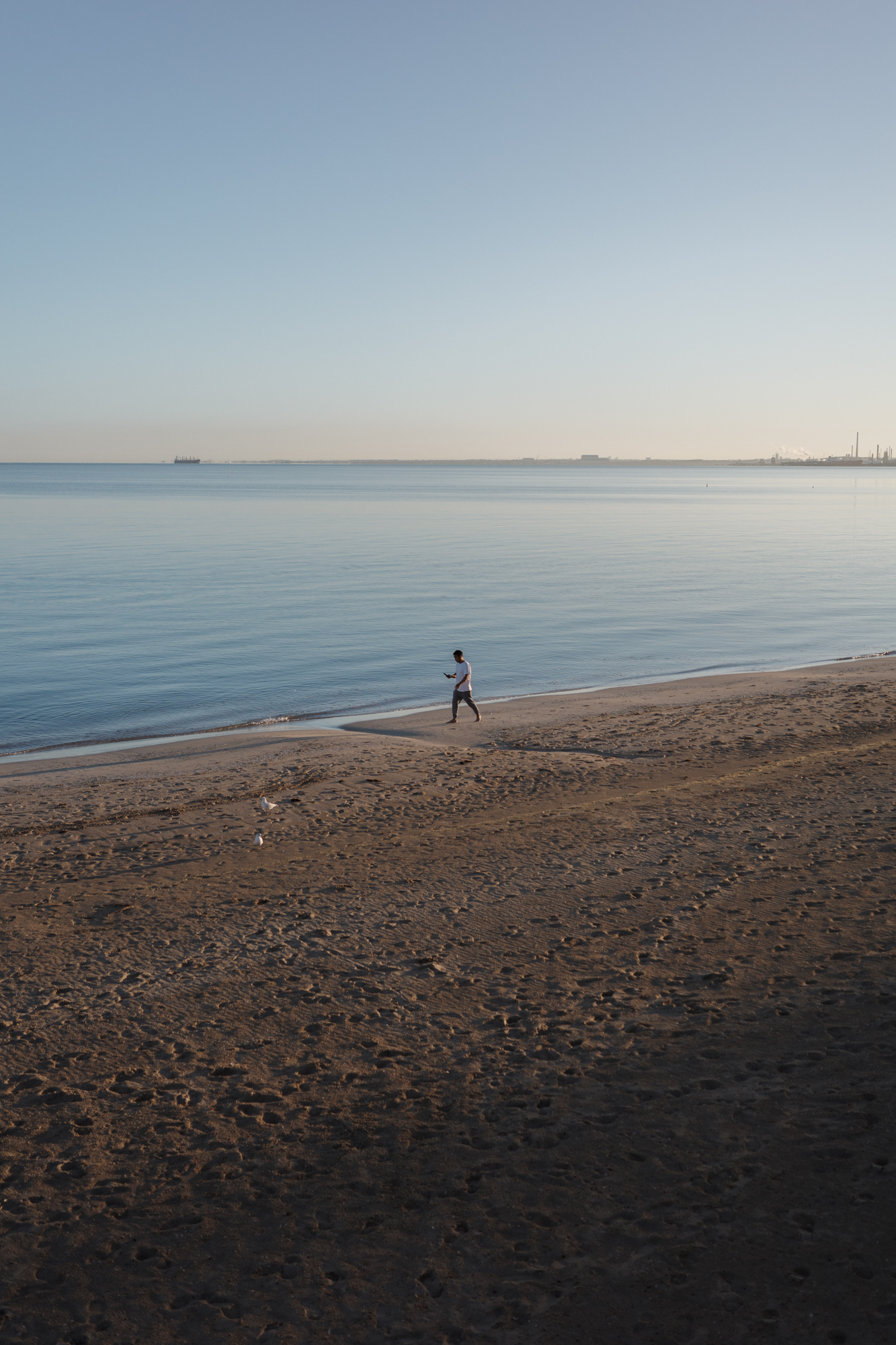 Auto-generated description: A lone individual jogs along the shoreline of a calm beach, with a clear sky above and the silhouettes of distant structures on the horizon.