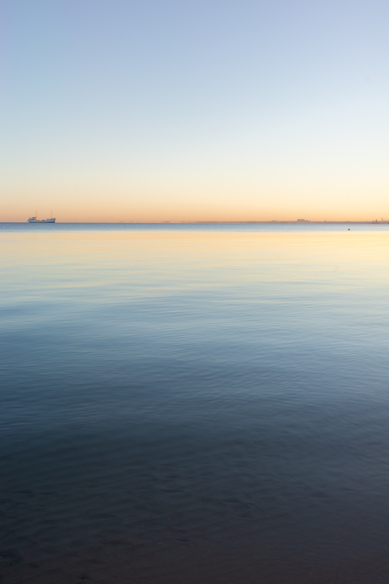 Auto-generated description: A calm sea under a gradient sunrise sky with a distant ship on the horizon.