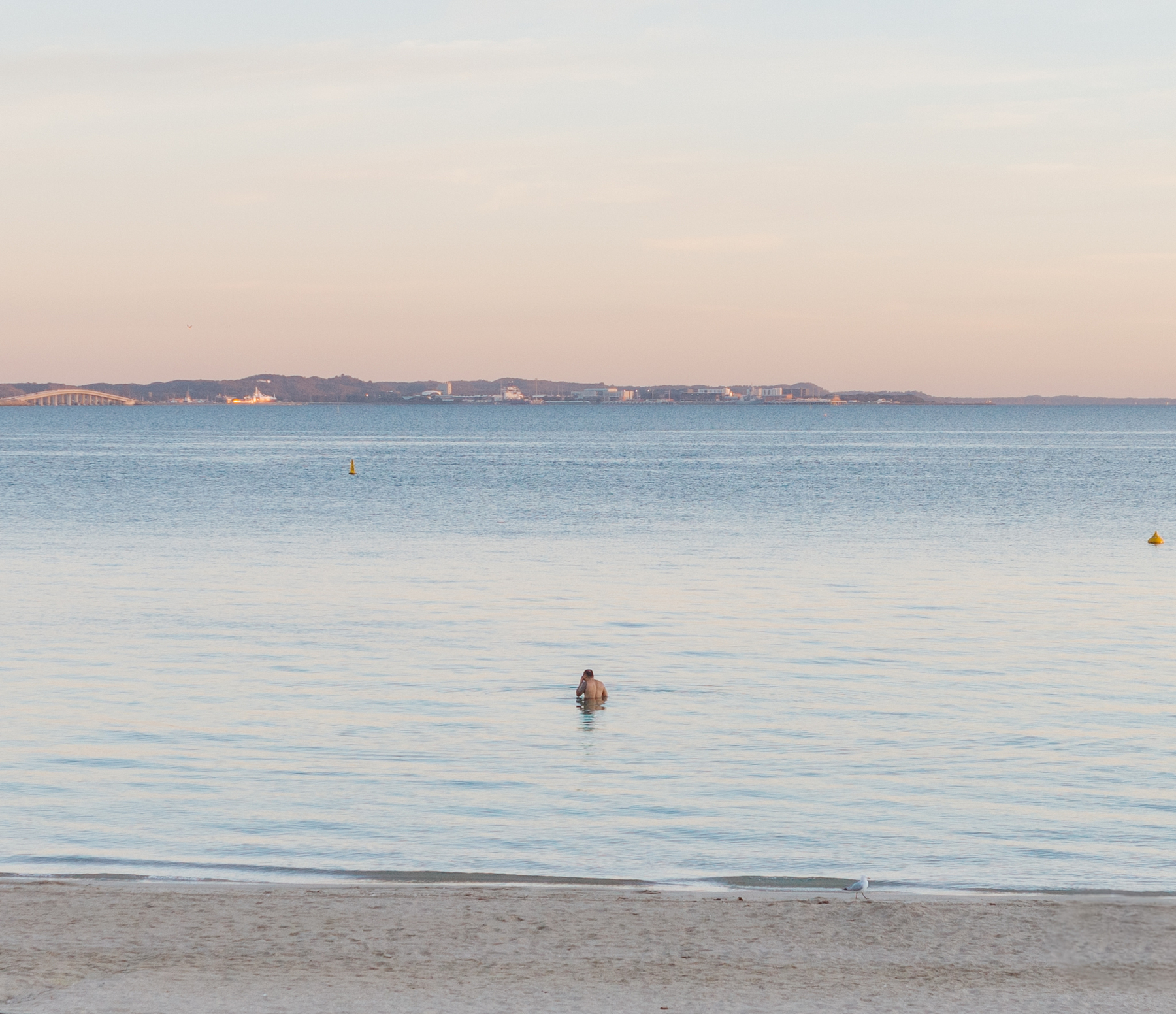 Auto-generated description: A solitary person is wading in calm sea waters near a sandy beach with a landmass visible on the horizon during what appears to be dusk or dawn.