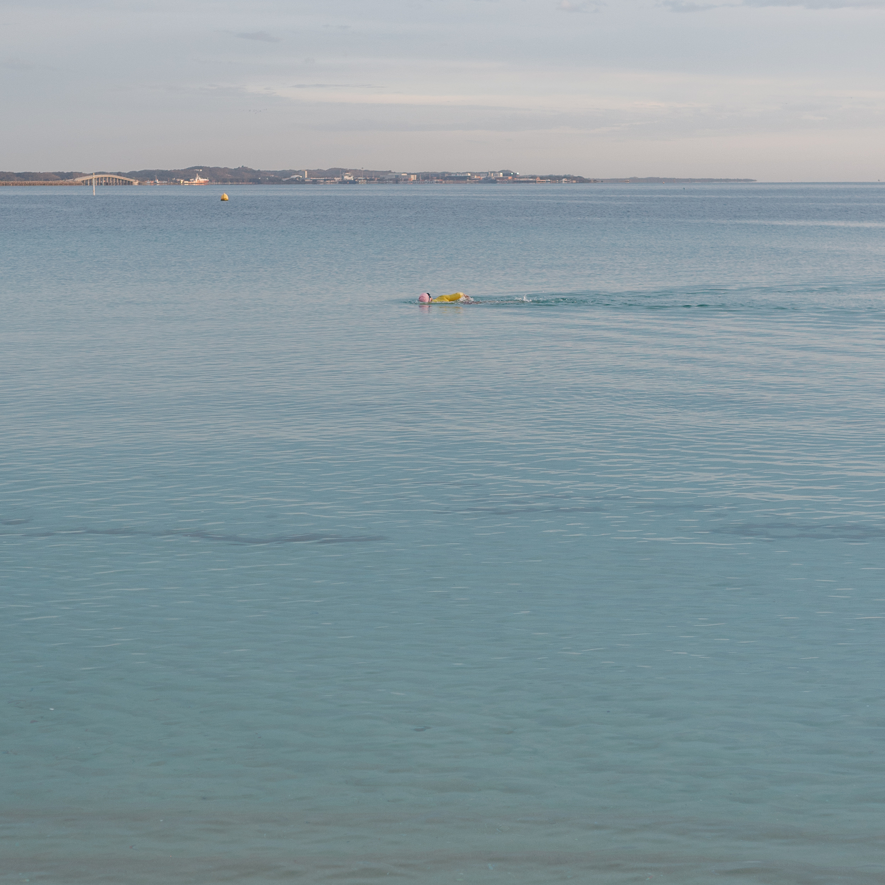 Auto-generated description: A person is swimming in a calm blue sea with a buoy nearby, and a bridge connects the distant landmasses under a soft cloudy sky.